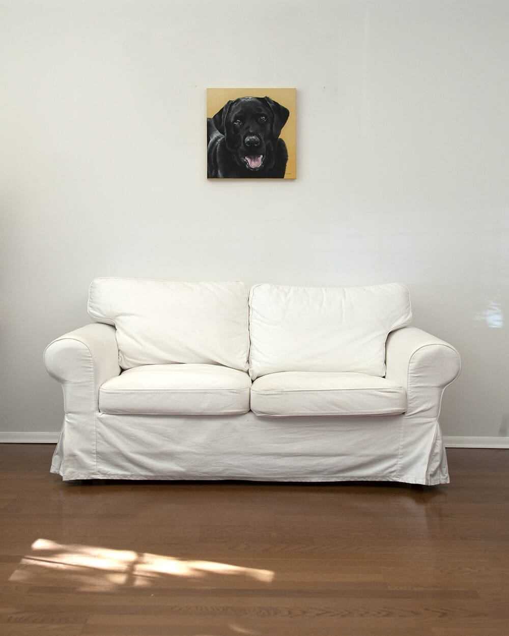 A custom dog portrait of a black labrador dog by fine arts painter Erica Eriksdotter hangs over a white loveseat