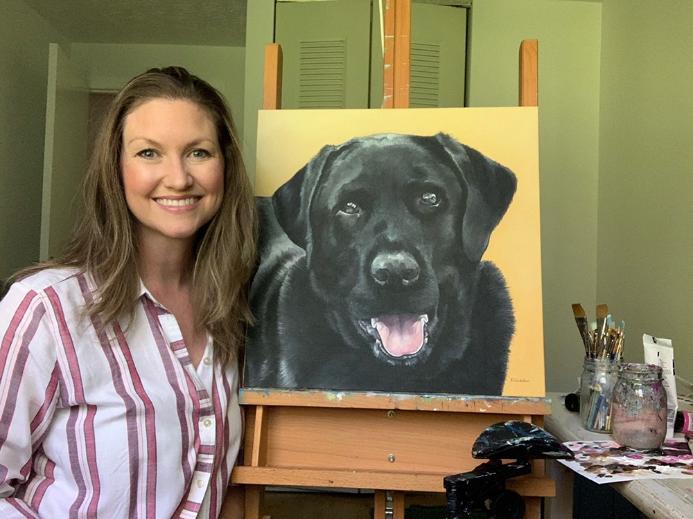 Fine arts painter Erica Eriksdotter sits by her easel with a custom painting of a black lab