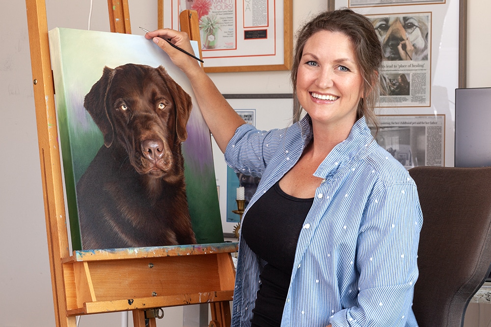 Artist Erica Eriksdotter painting a custom pet portrait of a brown Labrador in her studio, showcasing her detailed artwork and smiling while holding a brush at the easel.